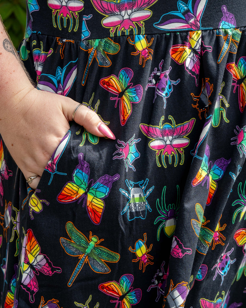 Close‑up of the Rebel Romance OUTLET PRIDE Bugs Stretch Smock Dress fabric. Bright rainbow bugs, dragonflies and beetles pop on black cotton, showing the bold alternative aesthetic and handy side pocket.