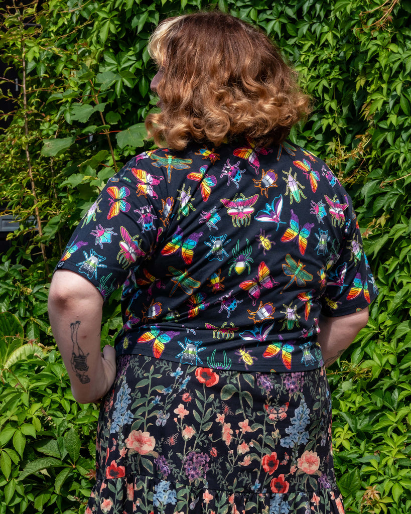 Back view of Maddie wearing the Rebel Romance OUTLET PRIDE Bugs Cropped Boxy Tee with bold rainbow insect prints, paired with a dark floral skirt. She stands outdoors against green foliage, showcasing ethical alternative fashion from a UK brand celebrating individuality.