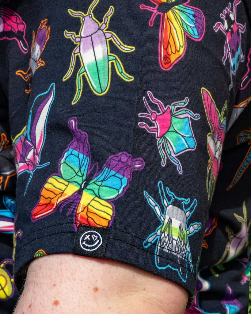Close‑up of the Rebel Romance OUTLET PRIDE Bugs Cropped Boxy Tee sleeve showing vibrant rainbow bugs, butterflies and beetles printed on soft black fabric. A small Rebel Romance smiley logo tab is visible, reflecting fun alternative style from this UK ethical fashion label.