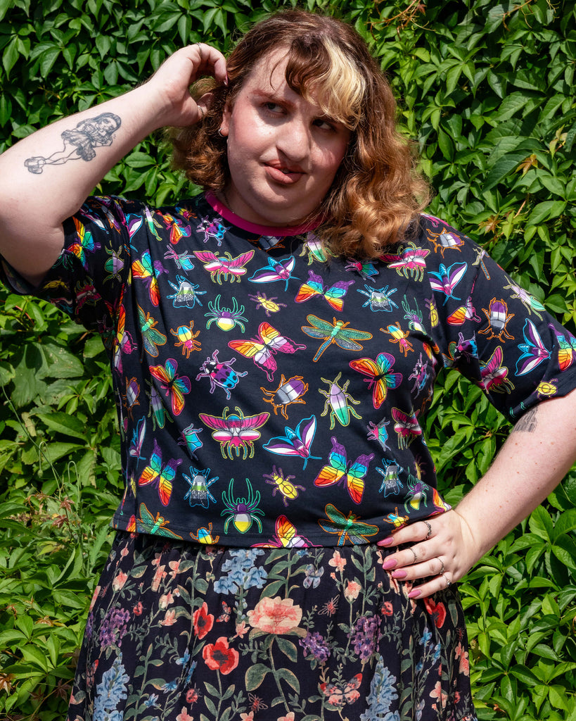 Maddie wears the Rebel Romance OUTLET PRIDE Bugs Cropped Boxy Tee with rainbow insect and bug illustrations, styled with a dark floral skirt. She poses outdoors against lush green leaves, celebrating bold alternative fashion from a small UK brand known for ethical slow fashion and vibrant designs.