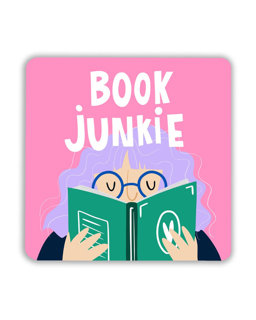 A square pink coaster featuring an illustration of a person with lilac curly hair and round blue glasses, holding a green book with a white leaf symbol. Above them, the words Book Junkie are written in white capital letters.