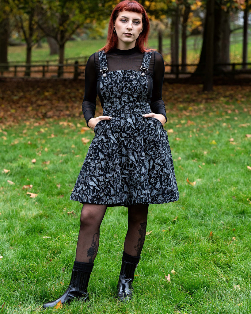 A person with red hair wears a black and grey illustrated flared pinafore dress over a sheer black long sleeve top. They hold the skirt out slightly to show the flare and look calmly toward the camera. They stand on green grass with autumn leaves scattered around.