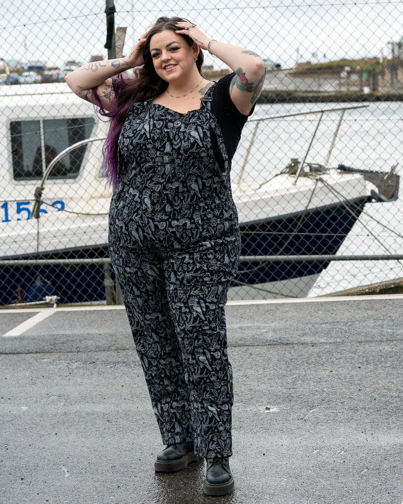 Luisa is wearing the Rebel Romance Seek print stretch twill dungarees in black and grey. She stands in a harbour car park in front of a moored boat, smiling with her hands in her hair. The dungarees feature an all over witchy illustration print with animals, crystals, herbs, feathers and mystical motifs.