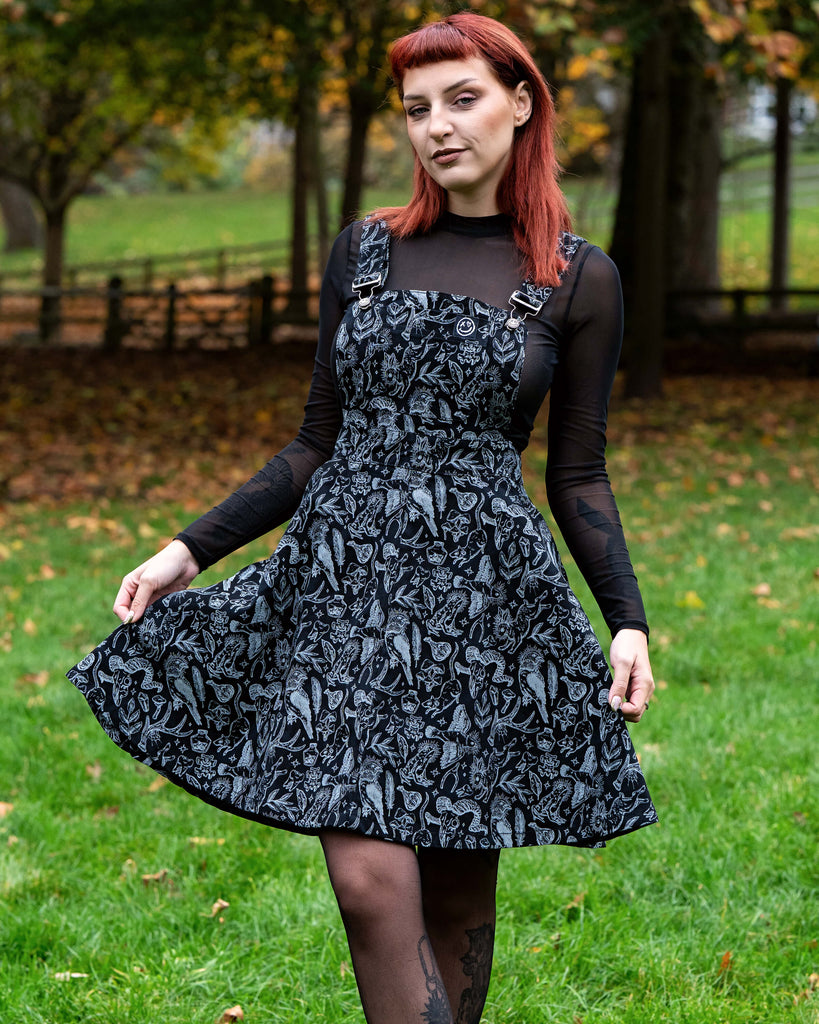 A person with chin length red hair stands on green grass wearing a black flared pinafore covered in grey mystical and nature inspired illustrations including birds, mushrooms and leaves. They layer it over a black sheer long sleeve top and wear black boots and patterned tights. They are smiling with hands behind their back and autumn trees are blurred behind them.
