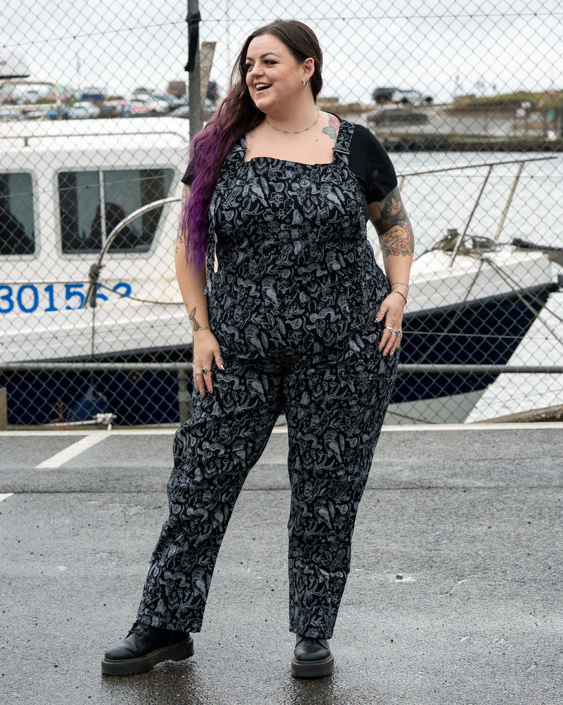 Luisa wears the Rebel Romance Seek print dungarees, standing outdoors in front of a docked boat. The black and grey mystical pattern covers the full outfit, featuring animals, skulls, herbs and magical symbols. She smiles and looks to the side, hands relaxed by her hips.