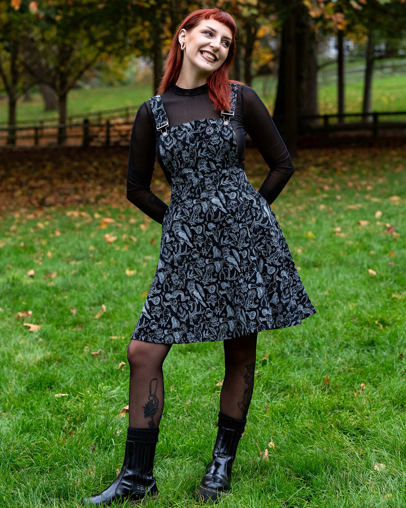 A person with red hair wears a black and grey illustrated flared pinafore dress over a sheer black long sleeve top. They hold the skirt out slightly to show the flare and look calmly toward the camera. They stand on green grass with autumn leaves scattered around.
