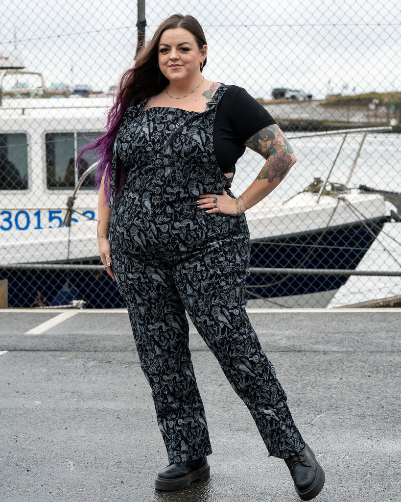 Luisa stands confidently in the Rebel Romance Seek print dungarees, facing the camera with her hand on her hip. The black and grey occult inspired print shows animals, crystals, plants and bones. A boat and harbour fencing sit in the background.