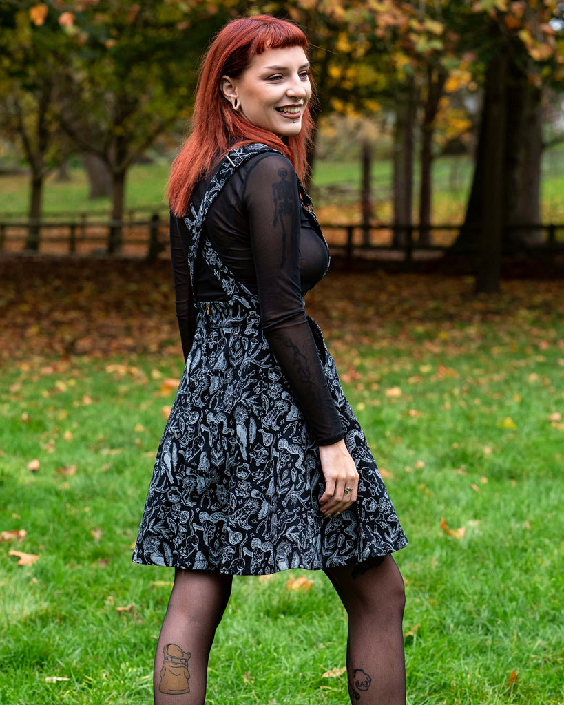 A person with red hair stands facing forward on grass wearing a flared pinafore with a grey illustrated print of animals, plants and magical motifs over a sheer black long sleeve top. They have hands in the pockets of the dress and wear black boots and patterned tights. Autumn trees and leaves sit behind them.