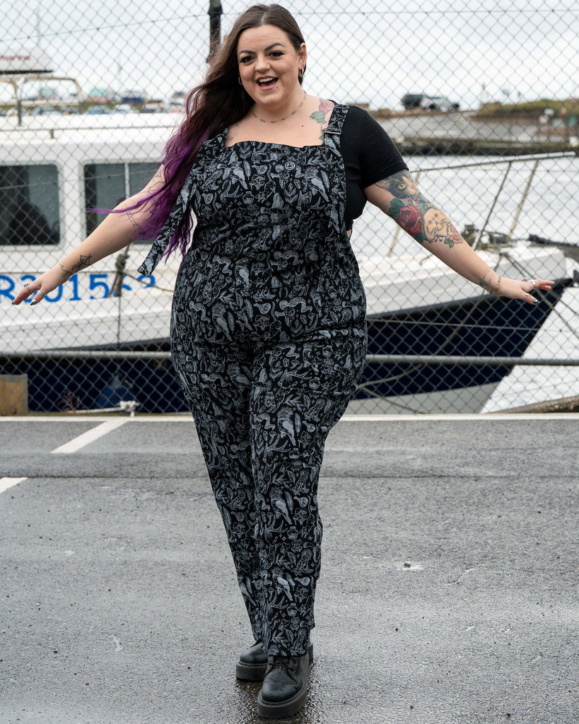 Luisa walks towards the camera wearing the Rebel Romance Seek print dungarees. She smiles with her arms open and the illustrated black and grey print shows magical animals, herbs, crystals and witchy motifs. A boat and harbour fencing are behind her.