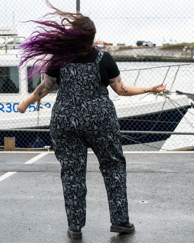 Back view of Luisa in the Rebel Romance Seek print dungarees. Her long purple hair blows in the wind while she stands in a harbour car park. The all over black and grey mystical print features animals, herbs and magical symbols.