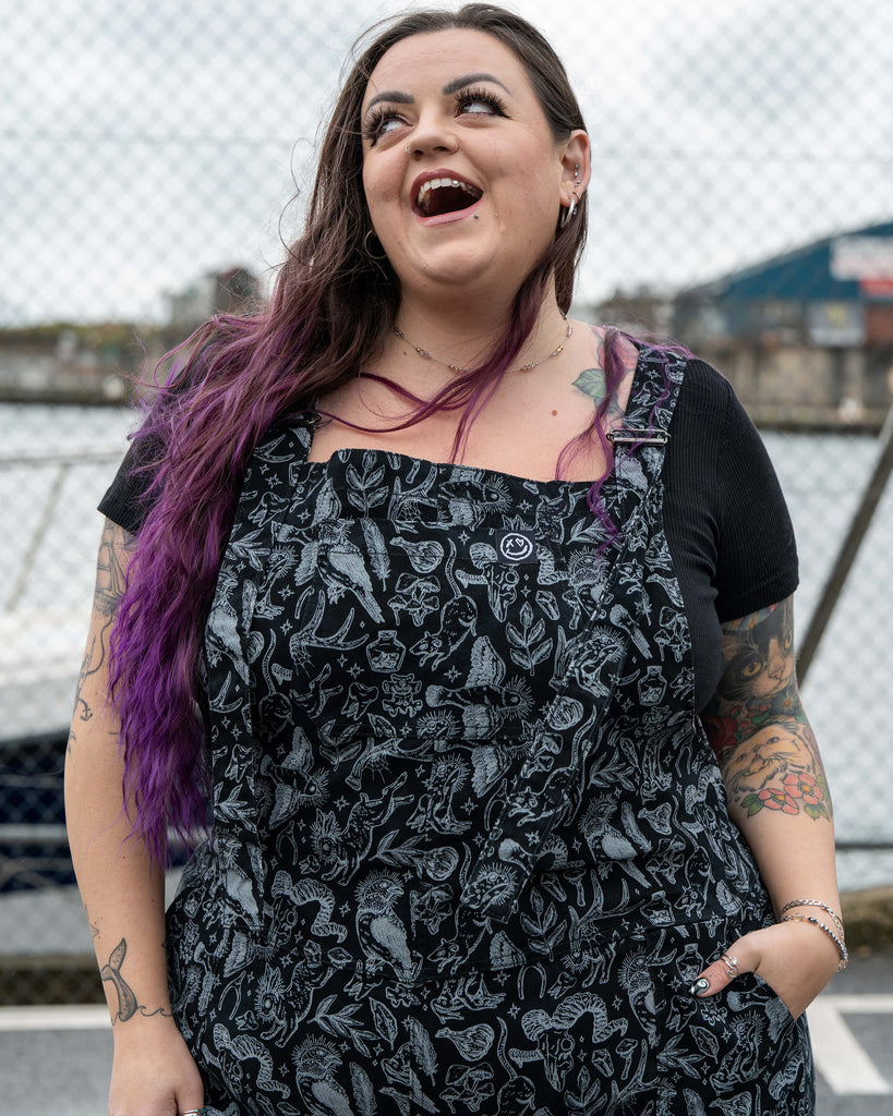 Close up detail of the Seek print fabric. The black and grey artwork features detailed illustrations of animals, feathers, crystals, mushrooms, herbs and magical motifs on the dungaree bib.
