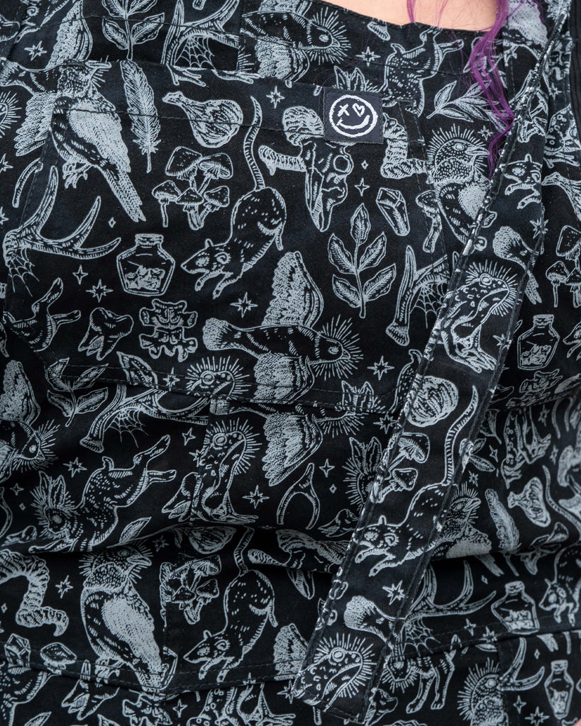 Close up of the hem and lower legs of the Rebel Romance Seek print dungarees worn by Luisa. The black boots and rolled cuffs show the illustrated mystical pattern with animals, plants and witchy icons.