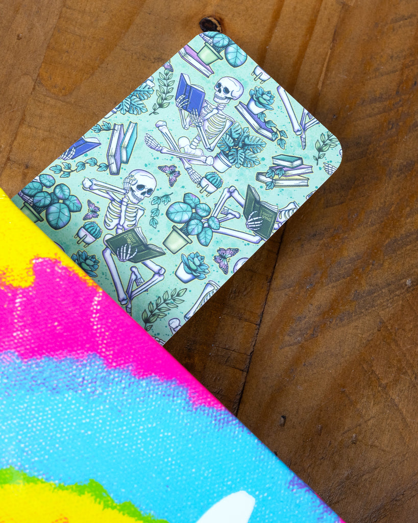A skeleton-themed Rebel Romance bookmark peeking out of a brightly coloured book, with a design of skeletons, books, plants, and butterflies on a green background.