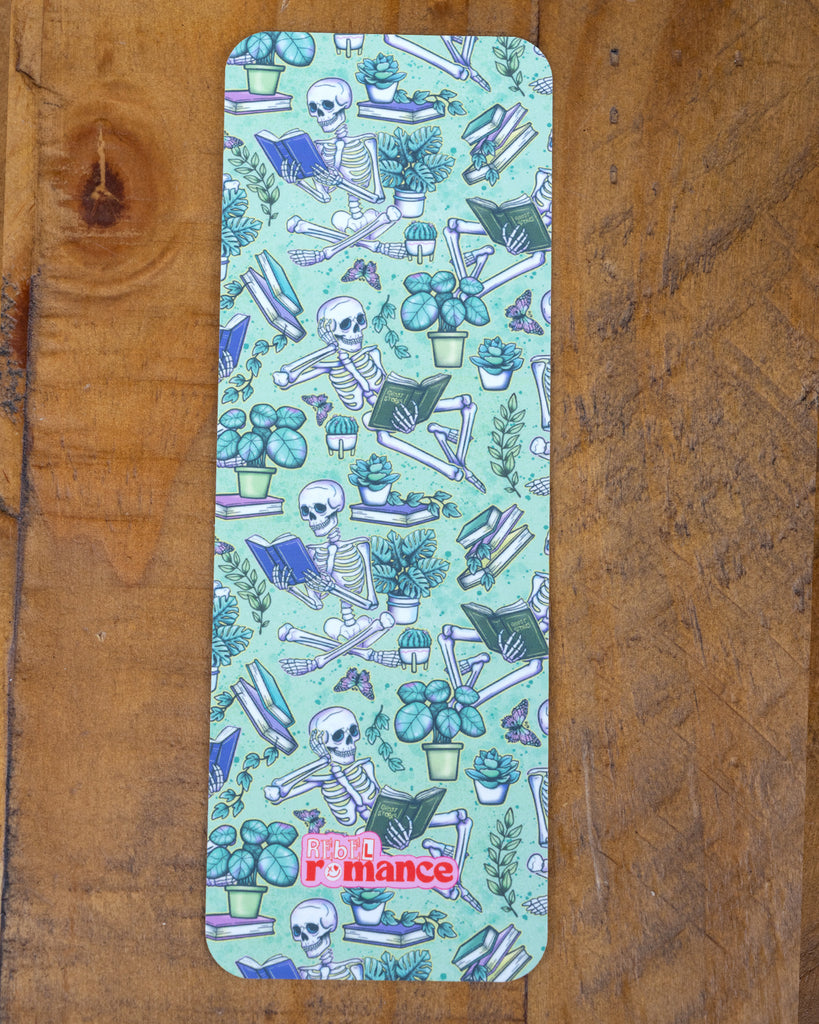 A close-up of the skeleton reading-themed Rebel Romance bookmark on a wooden surface, showing detailed illustrations of plants, butterflies, and bones on a mint background.