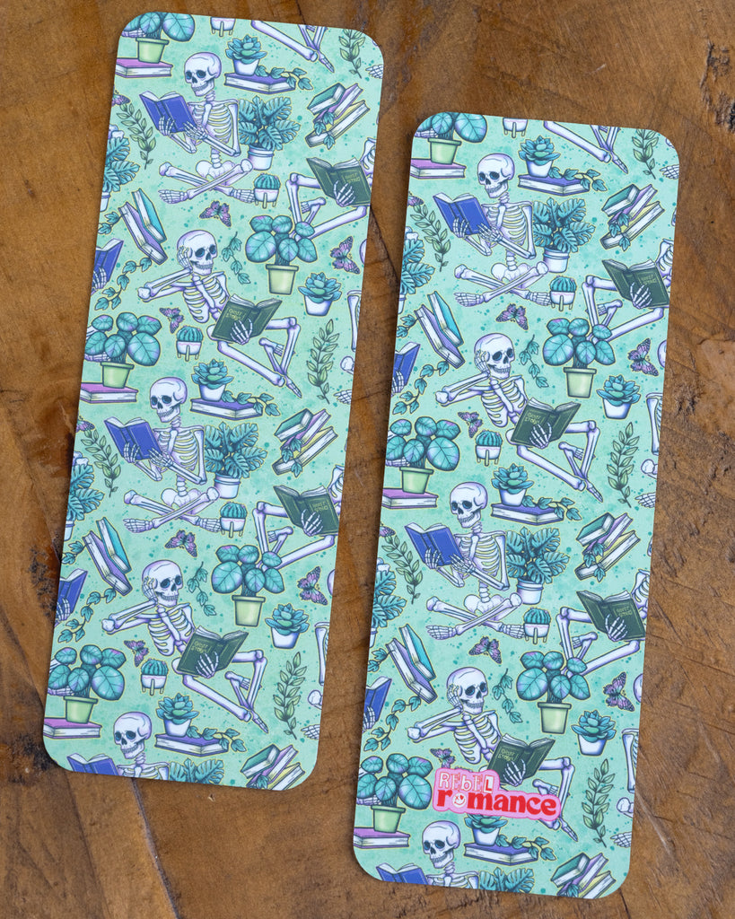 Two mint green Rebel Romance bookmarks on a wooden surface, both decorated with skeletons reading books among leafy plants and butterflies.