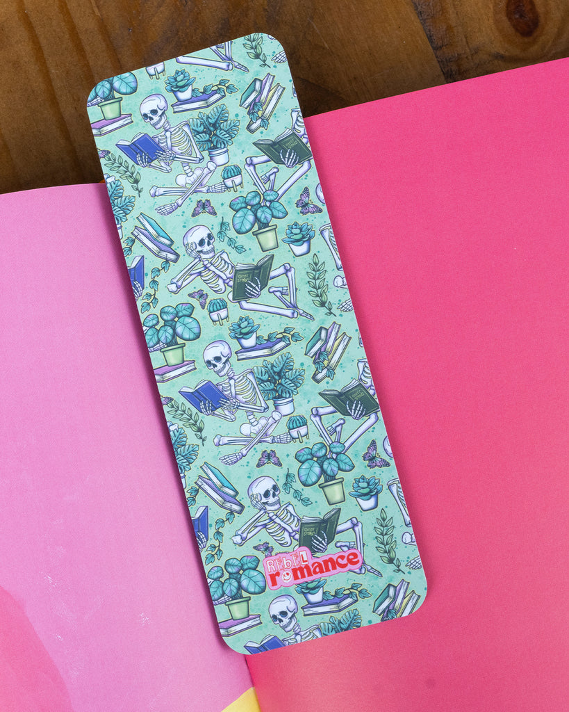 A close-up of the skeleton reading-themed Rebel Romance bookmark on a wooden surface, showing detailed illustrations of plants, butterflies, and bones on a mint background.