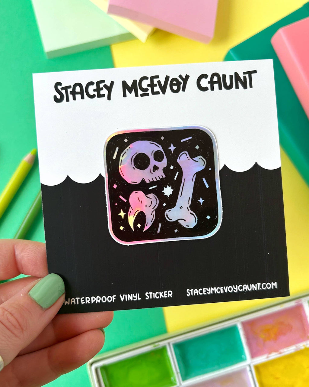 A square rainbow holographic sticker featuring a cartoon skull, bone, and tooth on a starry black background. The sticker is mounted on a branded backing card reading “Stacey McEvoy Caunt” with pastel stationery in the background.