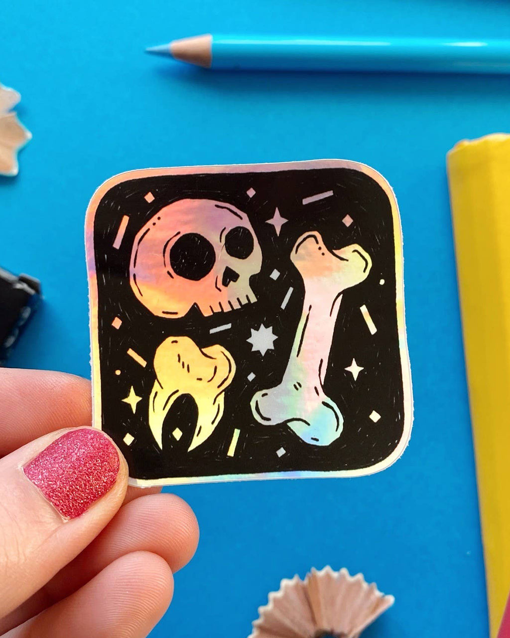  A close-up of the holographic sticker held by a hand with glittery red nails, showing its shiny rainbow surface. The sticker displays a skull, tooth, and bone with tiny stars around them.

