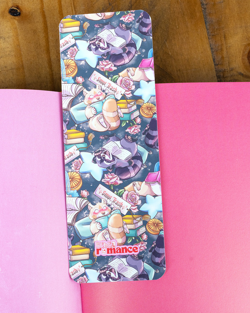  A close-up of a Rebel Romance Smut Trash bookmark placed inside a pink book, featuring a whimsical raccoon and red panda surrounded by books and celestial motifs