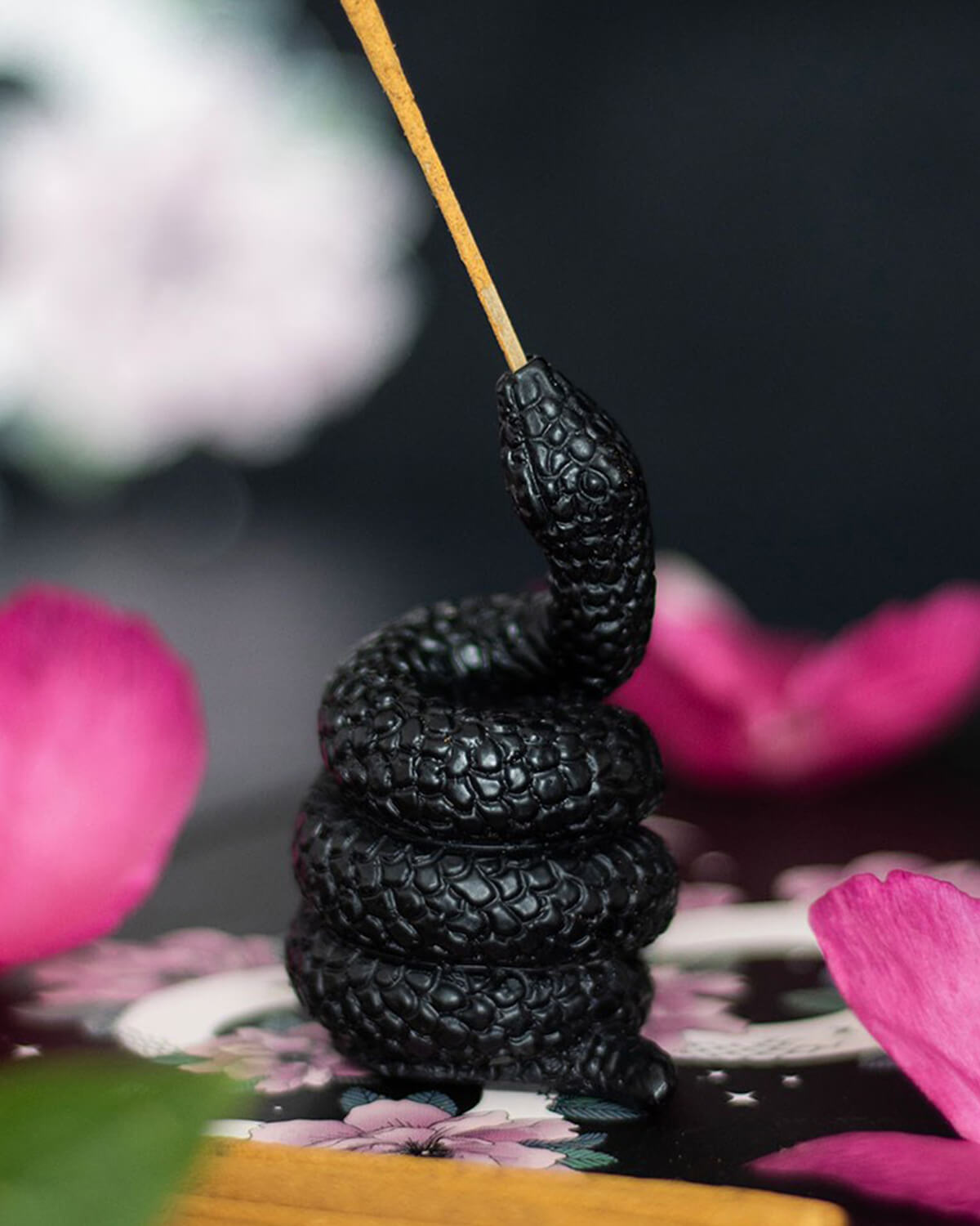 The black snake incense holder displayed on a floral background with pink flower petals and incense sticks, showcasing its textured details and glossy finish.
