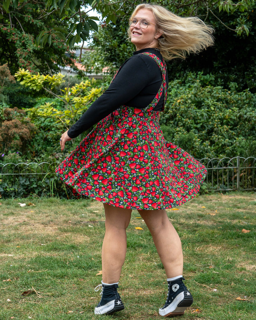 Model Betty twirls in the Rebel Romance Strawberry Field Flared Pinafore, showing the flared skirt detail against a garden backdrop.