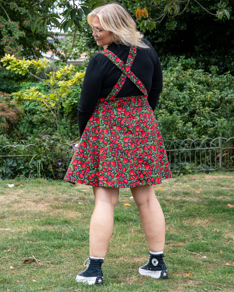 Betty wears the Rebel Romance Strawberry Field Flared Pinafore over a black long sleeve top, standing in a grassy park with trees in the background.