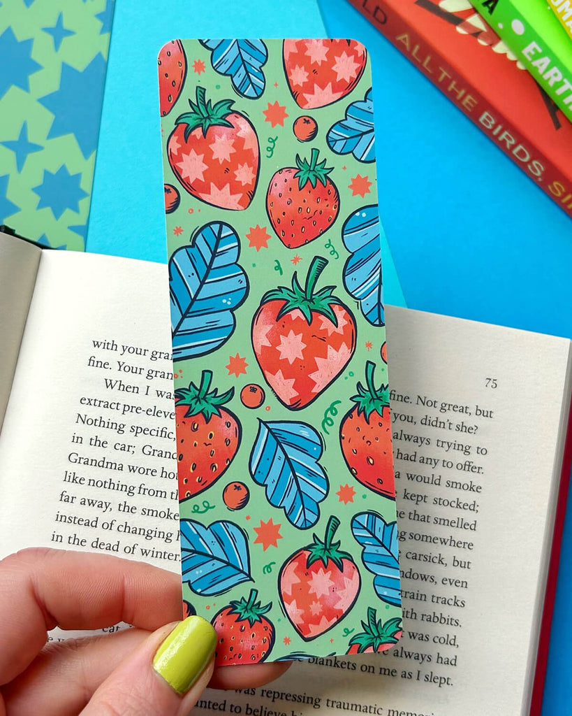  A hand with lime green nail polish holding a colourful bookmark with a pattern of illustrated red strawberries, blue leaves, and tiny stars on a soft green background. The bookmark rests on an open book with vibrant book covers stacked in the background.