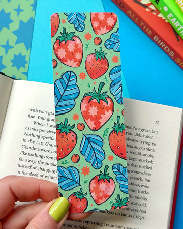 A hand with lime green nail polish holding a colourful bookmark with a pattern of illustrated red strawberries, blue leaves, and tiny stars on a soft green background. The bookmark rests on an open book with vibrant book covers stacked in the background.