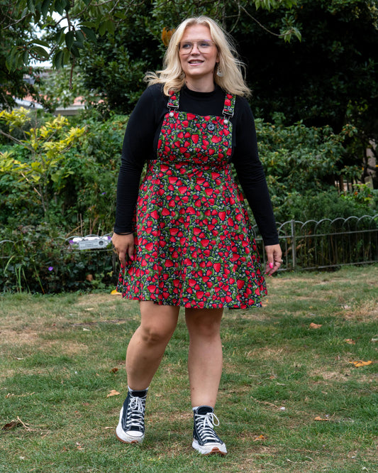 Betty wears the Rebel Romance Strawberry Field Flared Pinafore over a black long sleeve top, standing in a grassy park with trees in the background.