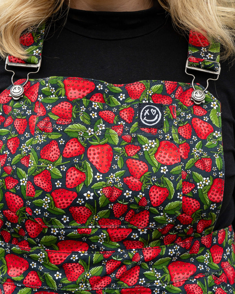 Fabric close-up of the Rebel Romance Strawberry Field Flared Pinafore, covered in bright red strawberries, green leaves, and white flowers.