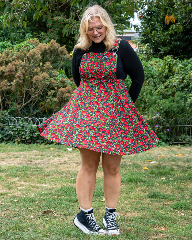 Model Betty shows off the Strawberry Field Flared Pinafore, styled with a neutral black top and white Converse trainers, featuring a vibrant fruit print.