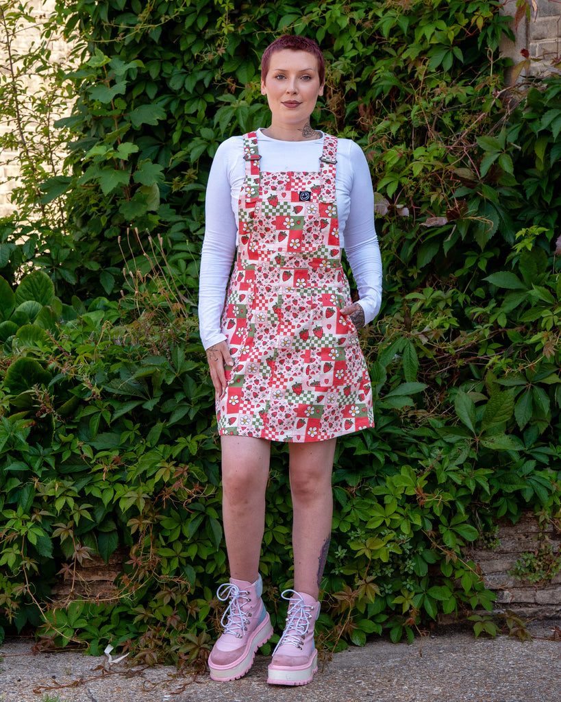 Femme model with short red purple hair wearing the rebel romance strawberry picnic pinafore with a white long sleeve top and pink platform boots. She is facing forward smiling with one hand in the pinafore dress pocket, stood in front of a lush green bush. The print on the pinafore is a sage green, pastel pink, pastel red and white colourblocks with strawberries and blossoms.