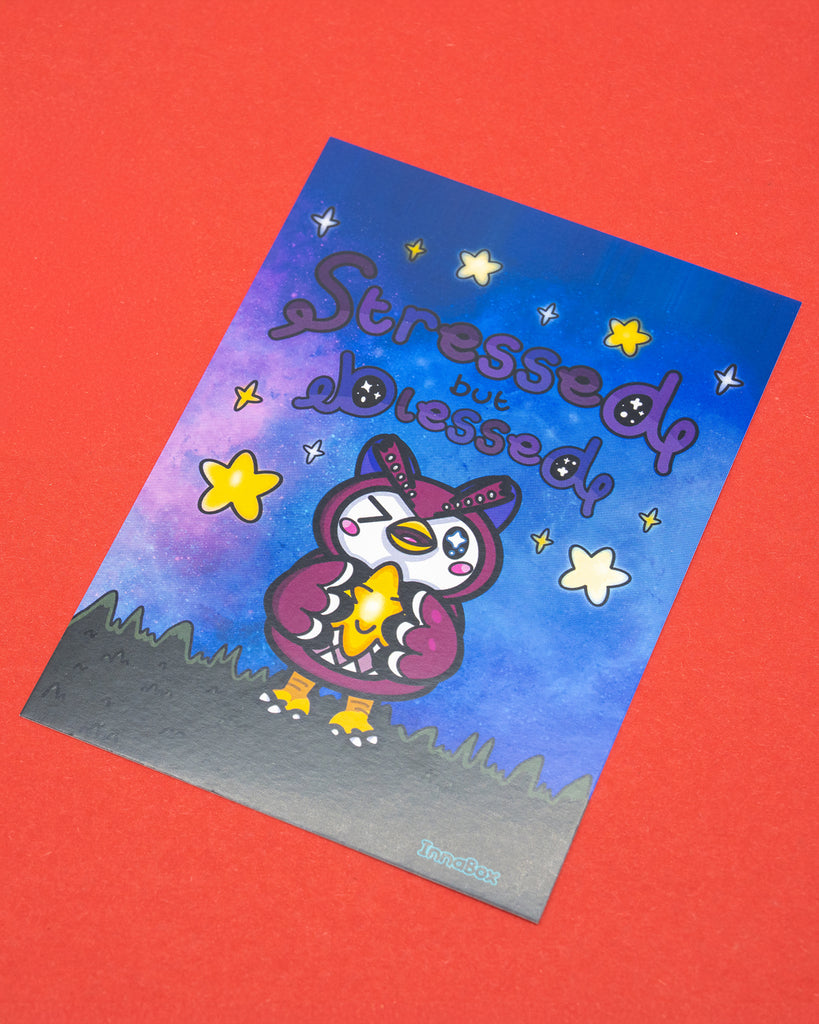A flat lay showing the Innabox Stressed But Blessed Celeste Postcard alongside its matching vinyl sticker. The postcard features a cute owl character holding a glowing star fragment against a dreamy purple and blue night sky with yellow stars. Printed on 350gsm silk board, standard UK postcard size, and blank on the reverse. A comforting disability inspired design perfect for gifting or displaying.