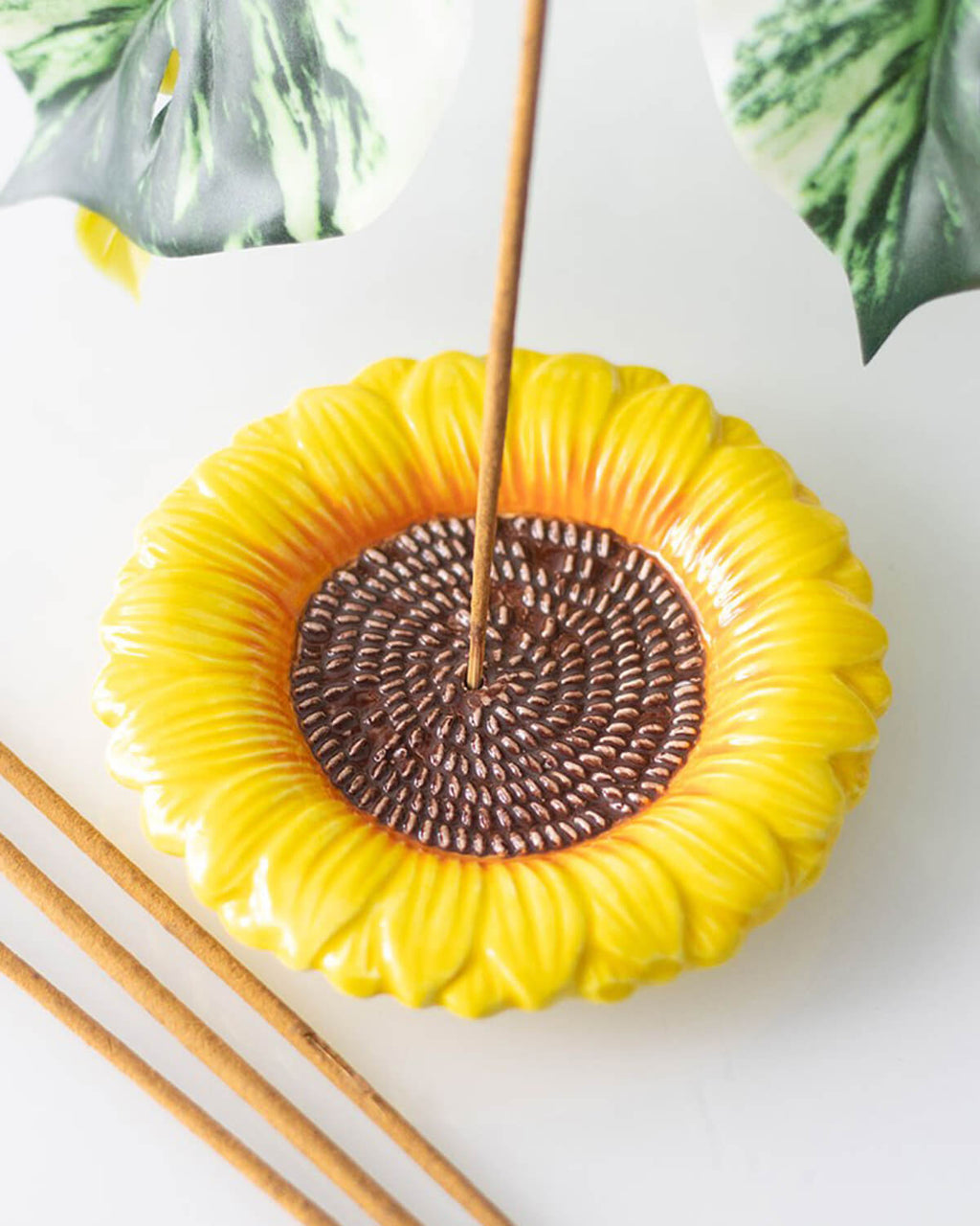Close-up view of the sunflower incense holder with an incense stick burning, placed on a white surface with green foliage in the background.