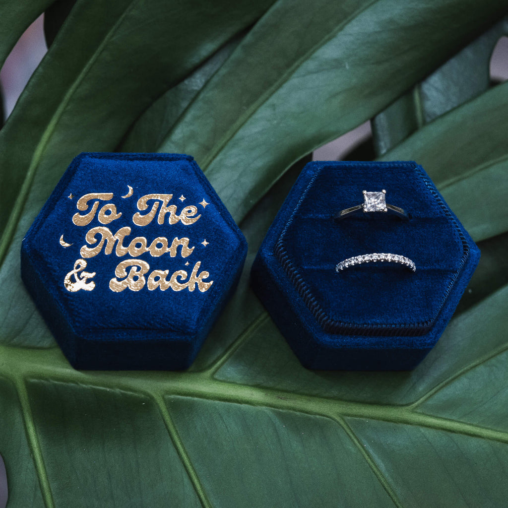 Two blue velvet hexagonal ring boxes, one showcasing gold-embossed “To The Moon & Back” and the other opened to reveal a sparkling engagement ring and wedding band, placed artistically on a green leafy surface.