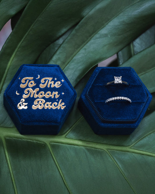Two blue velvet hexagonal ring boxes, one showcasing gold-embossed “To The Moon & Back” and the other opened to reveal a sparkling engagement ring and wedding band, placed artistically on a green leafy surface.
