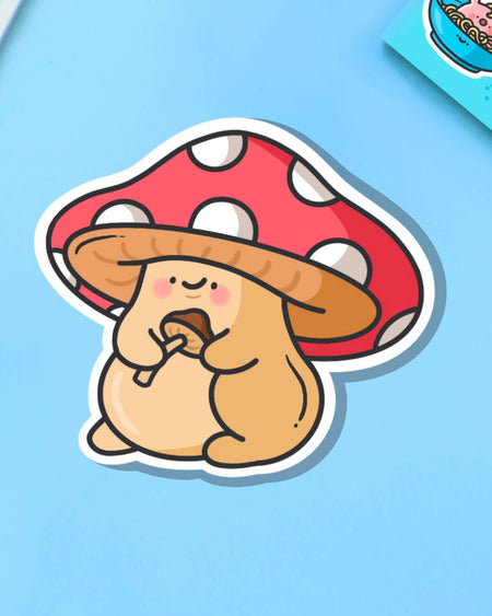 A cute cartoon-style vinyl sticker of a chubby tan mushroom character with a red and white spotted cap, holding a smaller mushroom in its hands. Displayed on a blue background beside a teacup and flower. Waterproof, glossy finish – ideal for decorating everyday items.
