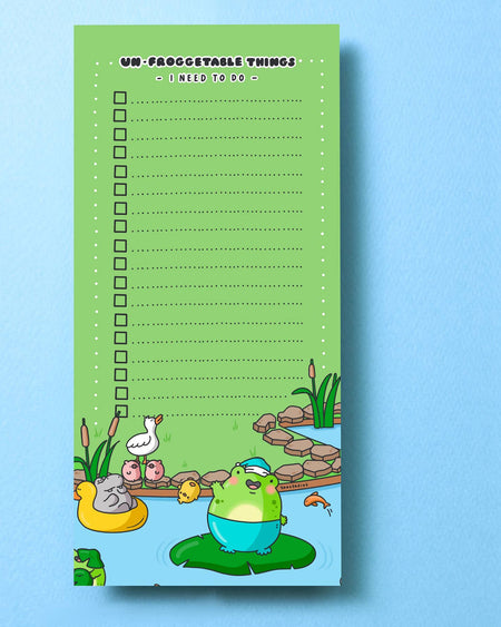 A green DL-sized to-do list pad titled “Un-froggetable things I need to do” with a checklist of dotted lines and tick boxes. The bottom features a cute cartoon frog in a blue floatie, sitting on a lily pad, surrounded by pond animals and plants. Perfect for fun and functional daily planning.