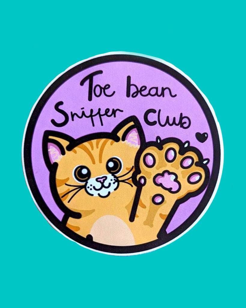 Cute round vinyl sticker featuring an illustrated orange tabby cat raising its paw with visible pink toe beans. Surrounded by a purple background and black outline, the sticker reads “Toe Bean Sniffer Club” in playful handwriting. Designed by Innabox and sold by Rebel Romance, a UK brand celebrating alternative style and millennial-friendly gifts.