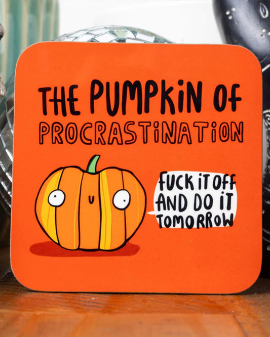 A bright orange coaster with rounded corners featuring a wide-eyed cartoon pumpkin character with the caption "The Pumpkin of Procrastination" in bold black lettering above it. A speech bubble from the pumpkin says, "F*ck it off and do it tomorrow." The coaster sits on a wooden surface with Halloween-themed decor in the background.