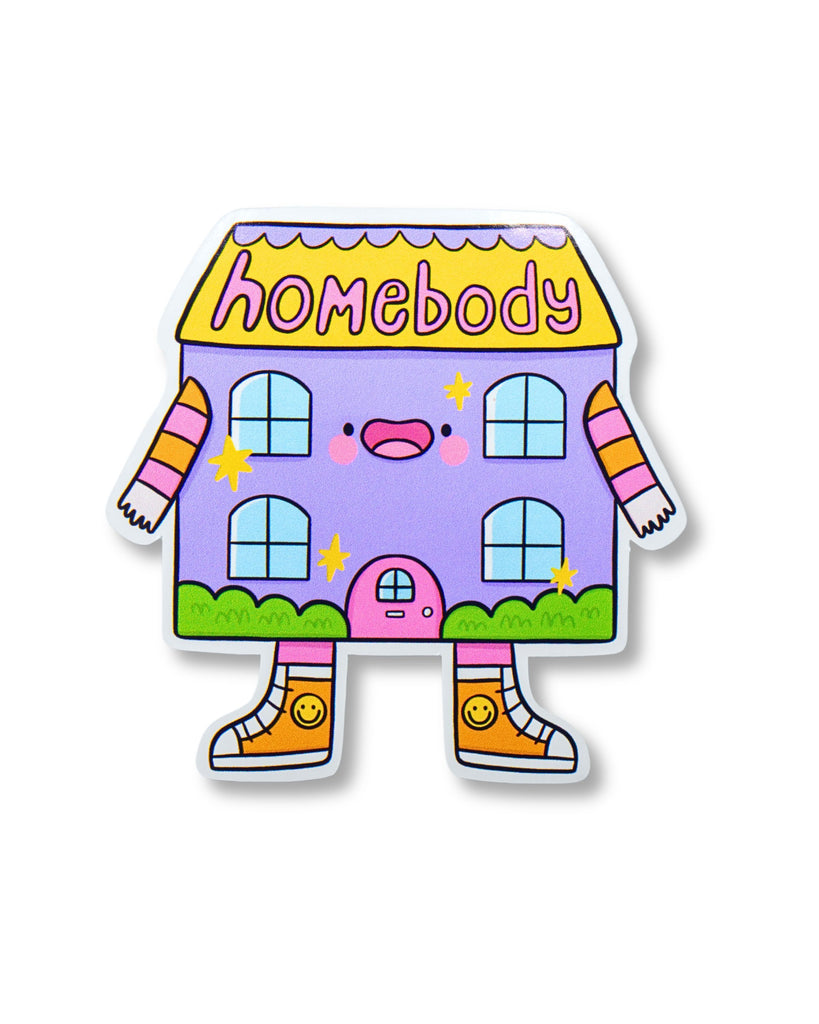 Cute illustrated glossy vinyl sticker shaped like a cartoon purple house with arms and legs wearing smiley face trainers. The word "homebody" is written on the yellow roof in playful text. Designed by Kellylou and sold by Rebel Romance, a small UK brand celebrating alternative fashion and cosy aesthetics. Ideal for introverts and lovers of staying in.