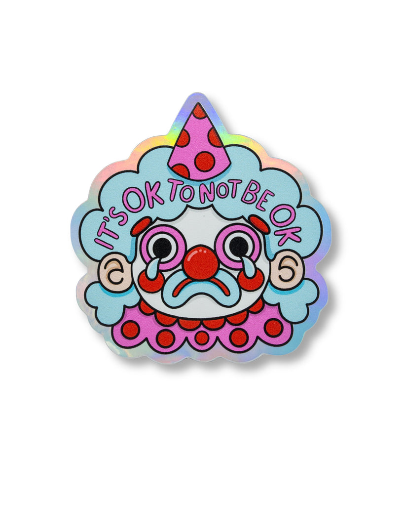 Holographic vinyl sticker featuring a sad clown with blue hair, a red nose, and a party hat, crying with the words “It’s OK To Not Be OK” above. Illustrated by Kellylou and sold by Rebel Romance, a small UK brand celebrating alternative fashion, style, and gifts for millennials. The sticker has a holographic matte finish and measures approximately 70mm.