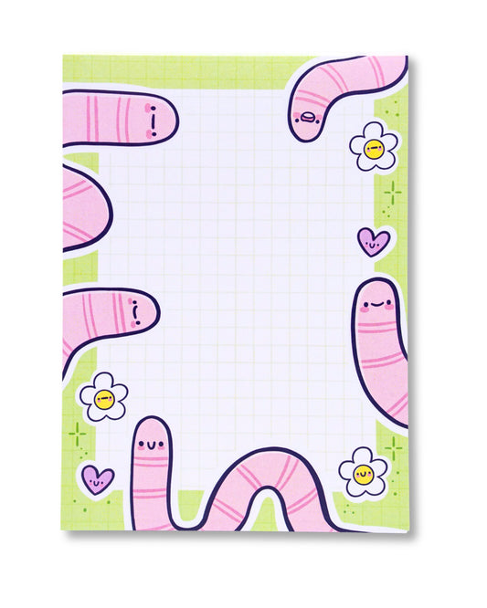 Unpackaged Spring Worms A6 Notepad by Kellylou, sold by Rebel Romance. The design includes pastel pink cartoon worms with smiley faces, cheerful flowers, and hearts along the border of a green grid layout. A fun and whimsical stationery pad perfect for everyday note-taking.