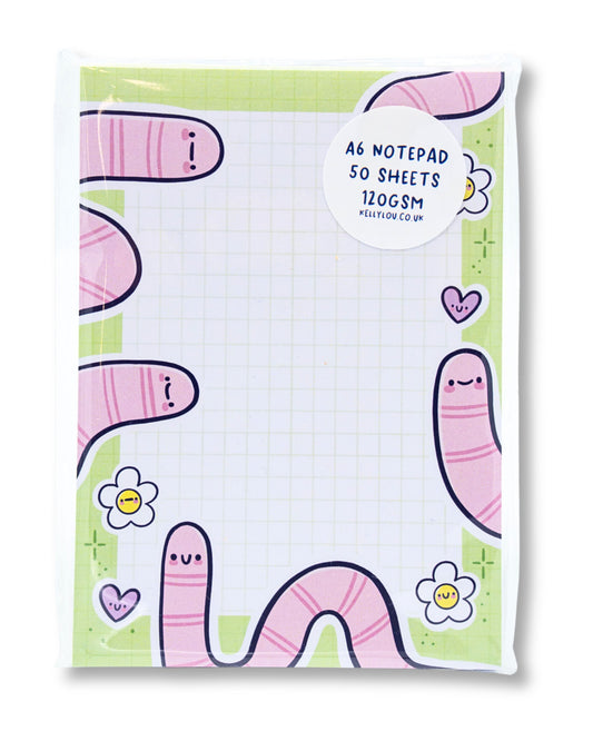 Photo of the Rebel Romance Spring Worms A6 Notepad by Kellylou, packaged in a clear wrap with a circular sticker showing product details. The notepad features a grid layout with cute pink worms, smiling flowers, and pastel hearts on a green border. Ideal for jotting down lists or notes with quirky charm.
