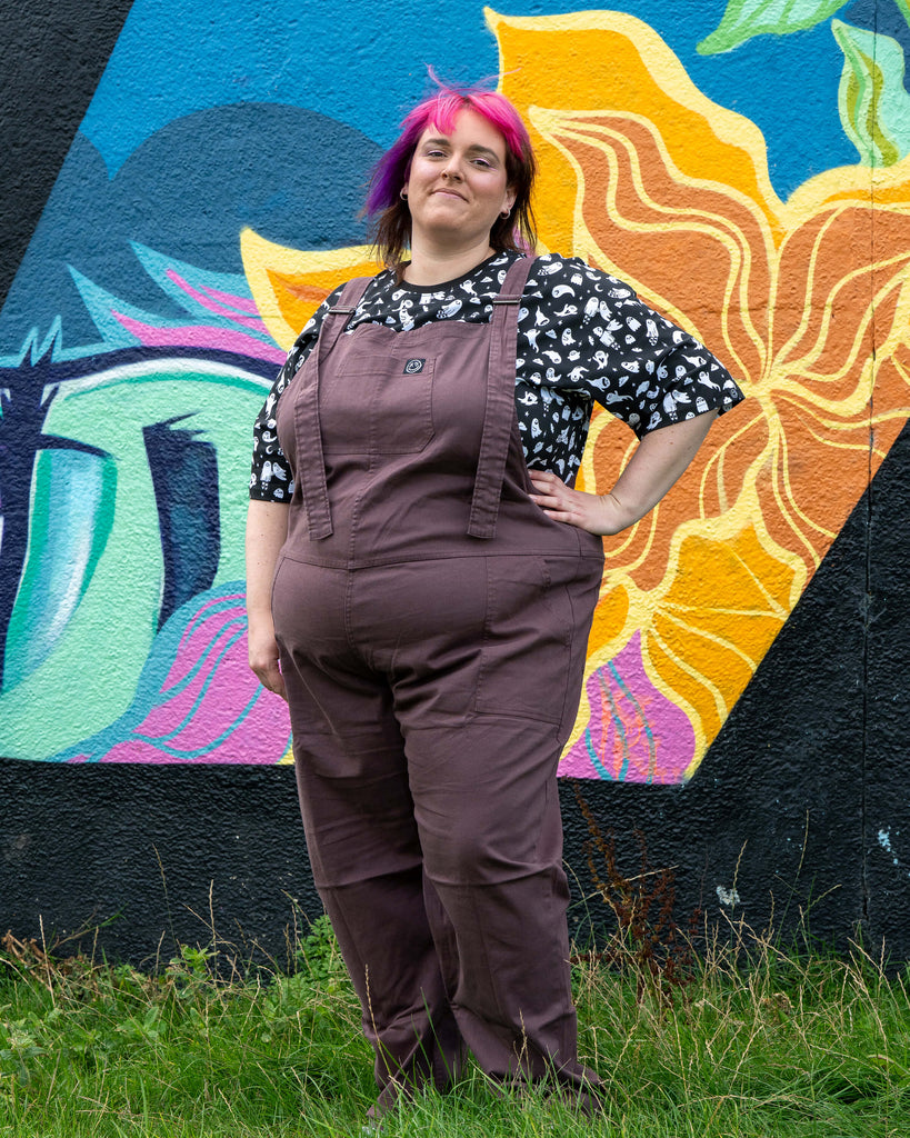 Full body shot of Chloe in the Plum Mocha dungarees, smiling with one hand on hip, mural in the background.
