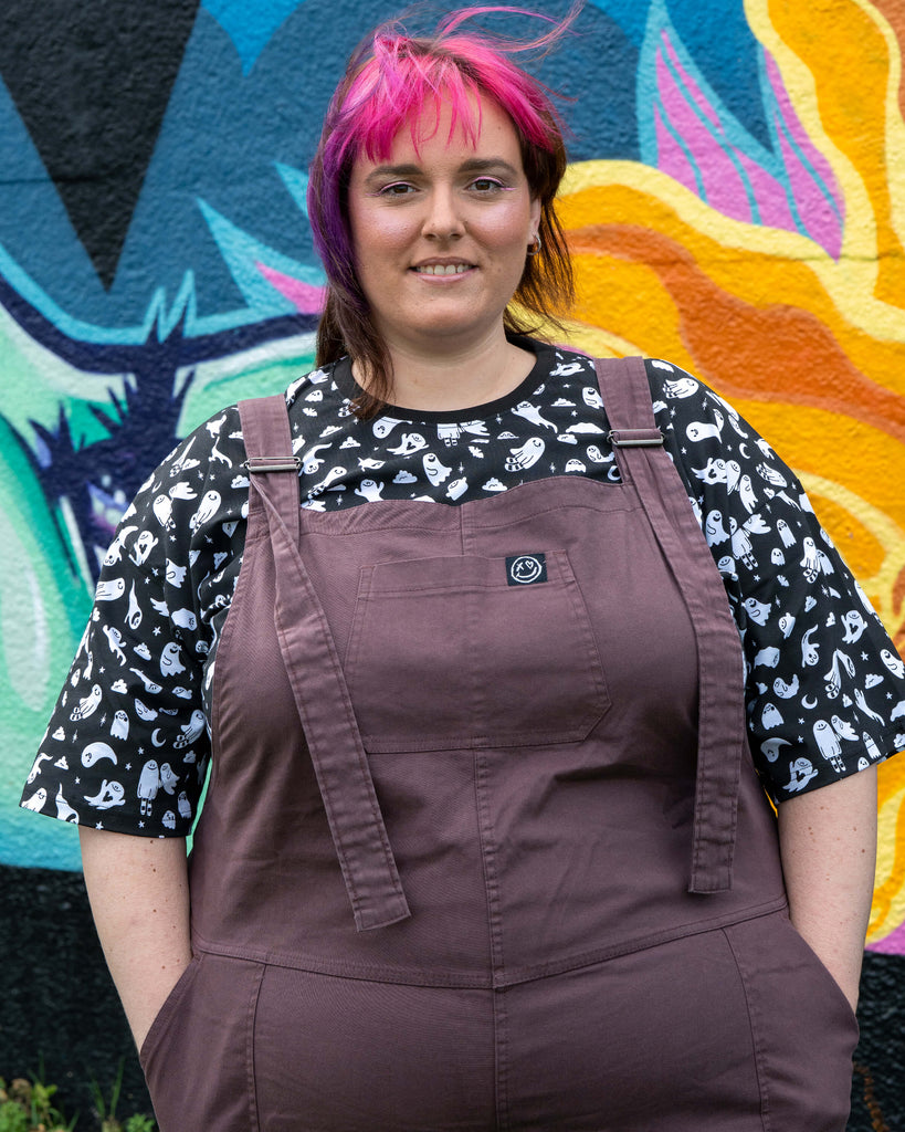 Chloe with pink and purple hair wears Rebel Romance Plum Mocha Stretch Twill Dungarees over a black and white ghost print t-shirt, standing in front of a colourful graffiti mural.