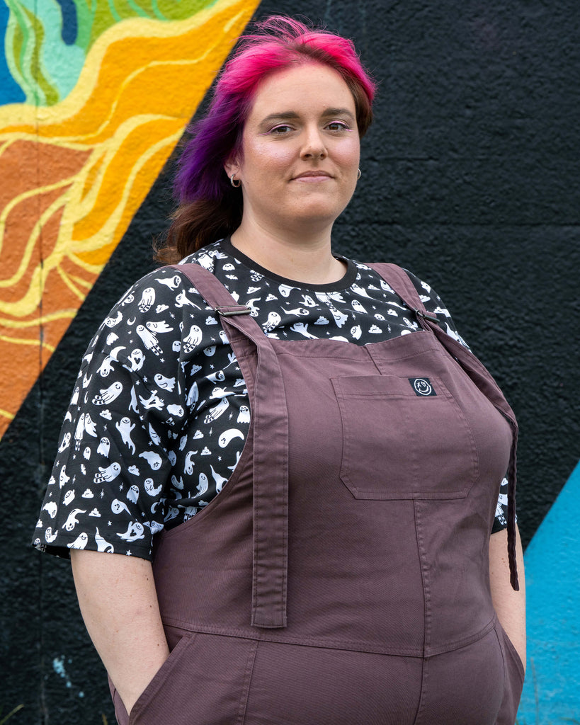 Side view of Chloe in the Plum Mocha Stretch Twill Dungarees with hands in pockets, showing the ghost print t-shirt underneath.