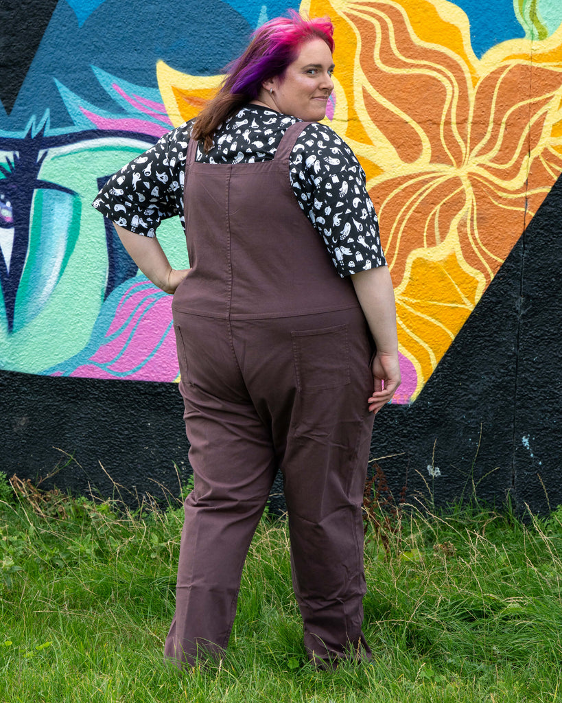 Back view of Chloe wearing the Plum Mocha dungarees, hand on hip, standing in front of the mural.