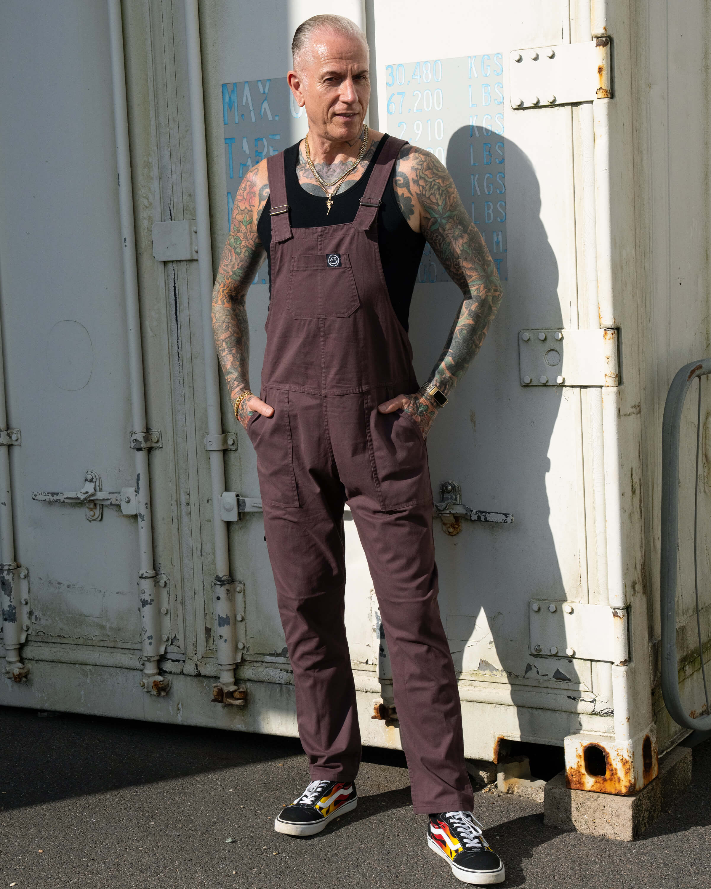 James with tattoo sleeves wears Rebel Romance Plum Mocha Stretch Twill Dungarees over a black vest, standing in sunlight against a white shipping container.