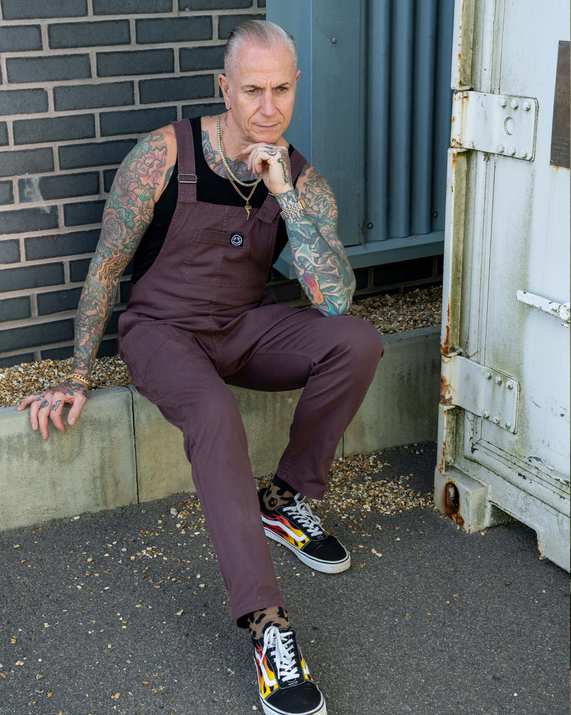 James sits on a kerb wearing the Plum Mocha dungarees, black vest, and patterned trainers, leaning casually.